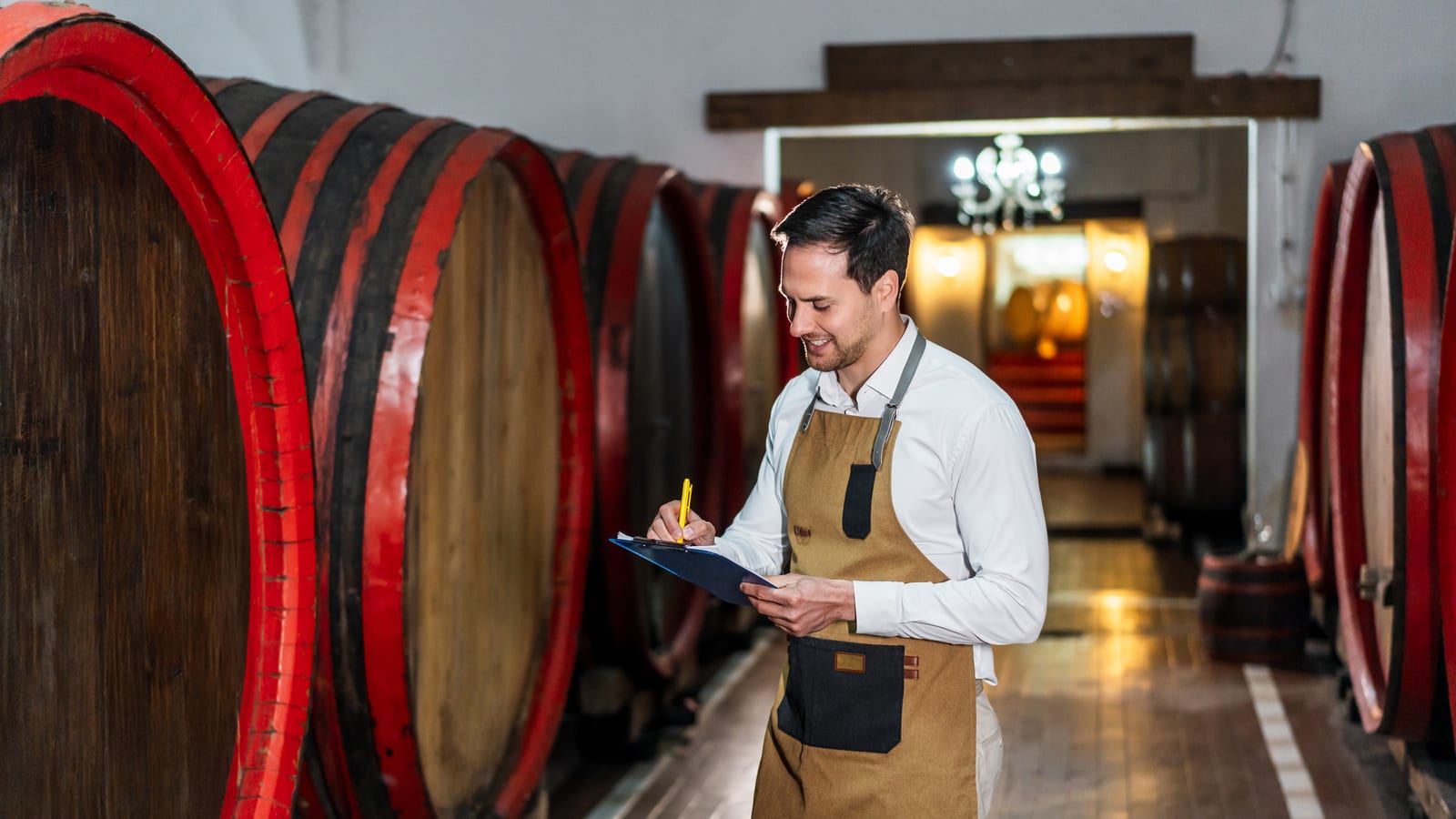 A smiling winemaker in a leather apron taking notes on a clipboard among red-ringed oak barrels in the cellar
